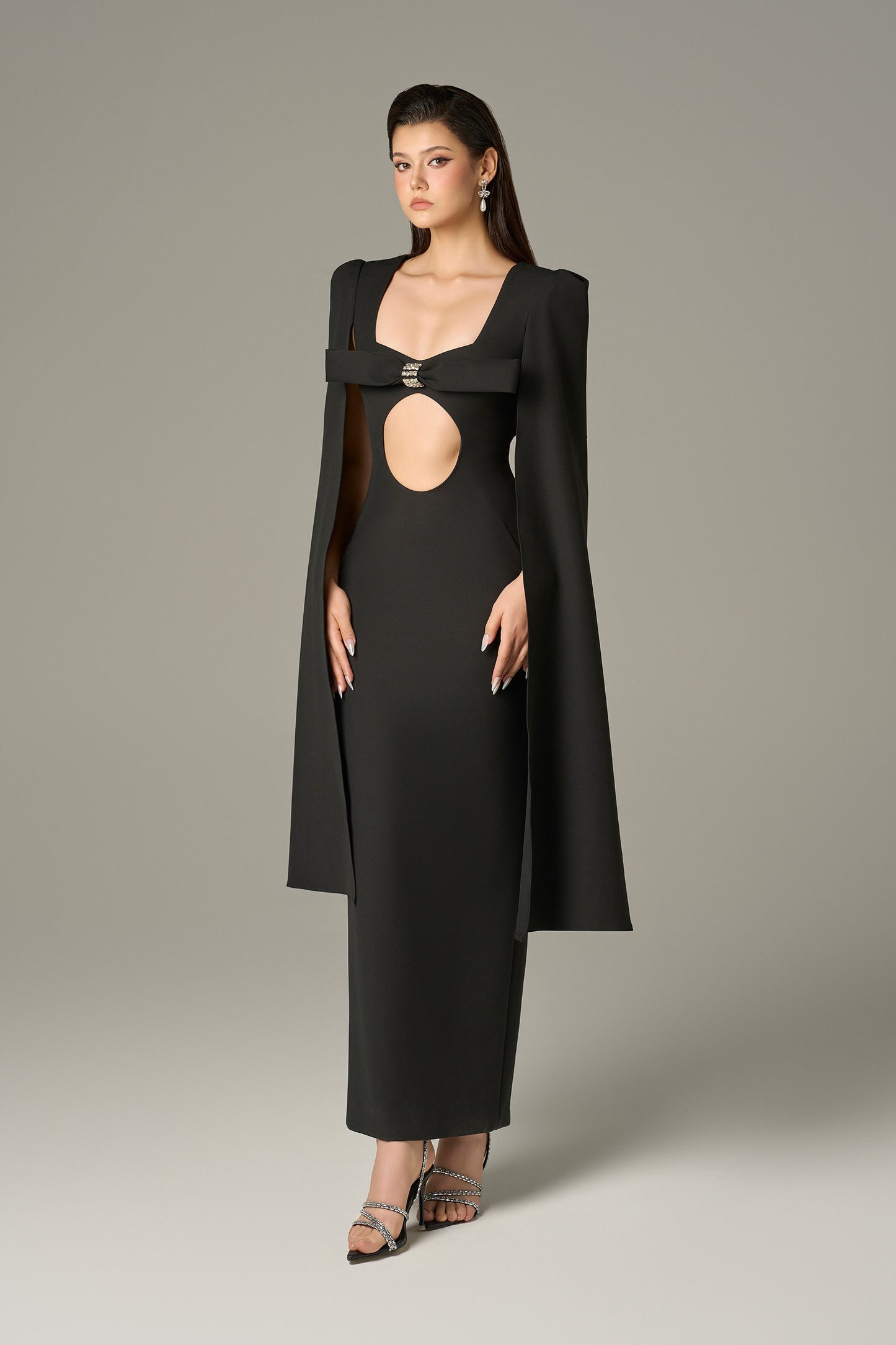 The Black Maxi Dress stuns with a crystal-embellished bow, bold circular cutout and flowing cape sleeves.
