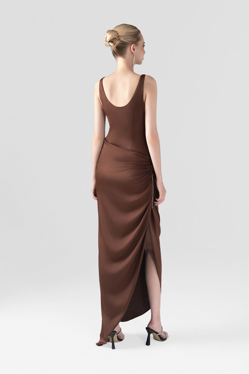 The brown silk maxi dress, detailed with a scoop neckline, hand-sewn rose accent, and stone chain embellishments.