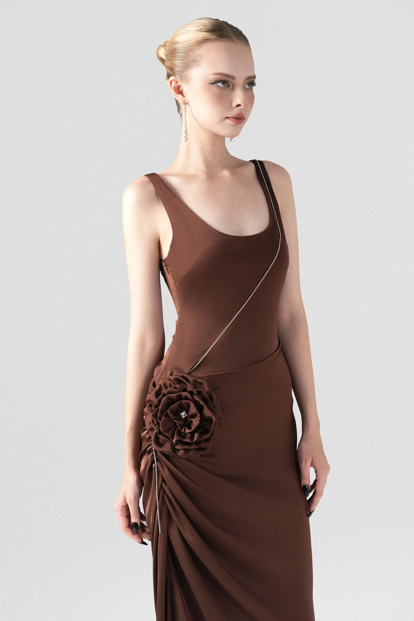 The brown silk maxi dress, detailed with a scoop neckline, hand-sewn rose accent, and stone chain embellishments.