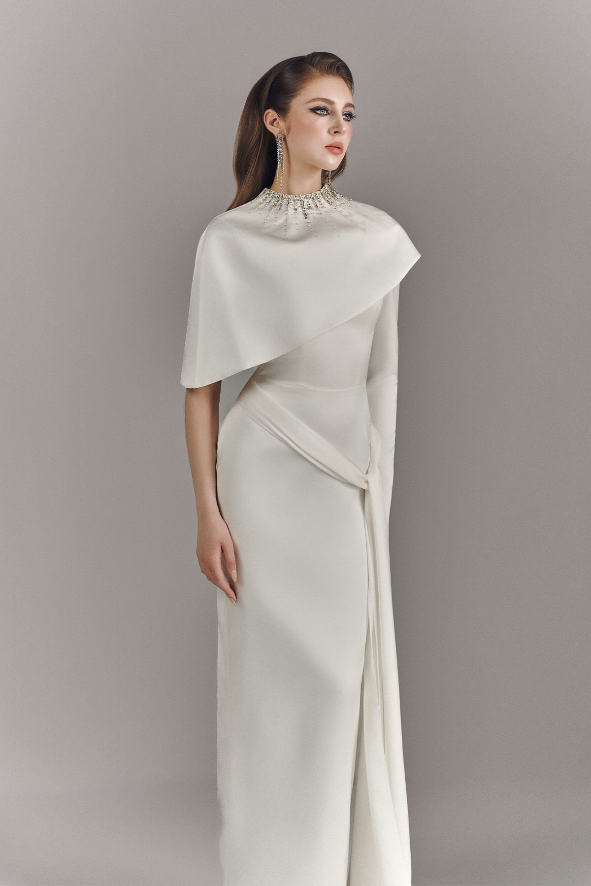 Woman wearing a long, elegant white dress with a cape against a plain background