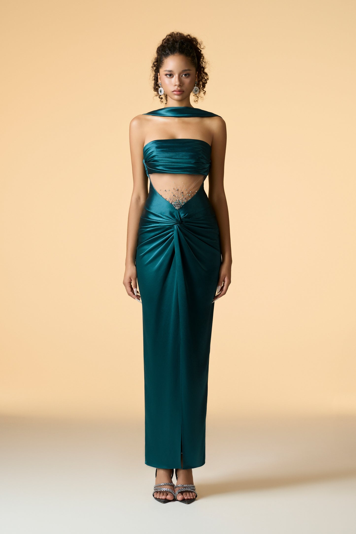 A sleeveless maxi dress in a dark, shimmering fabric, featuring a bandeau neckline with a scarf neck wrap. The dress has a draped front, a mesh waist, and hand-embellished crystals accentuating the bodice.