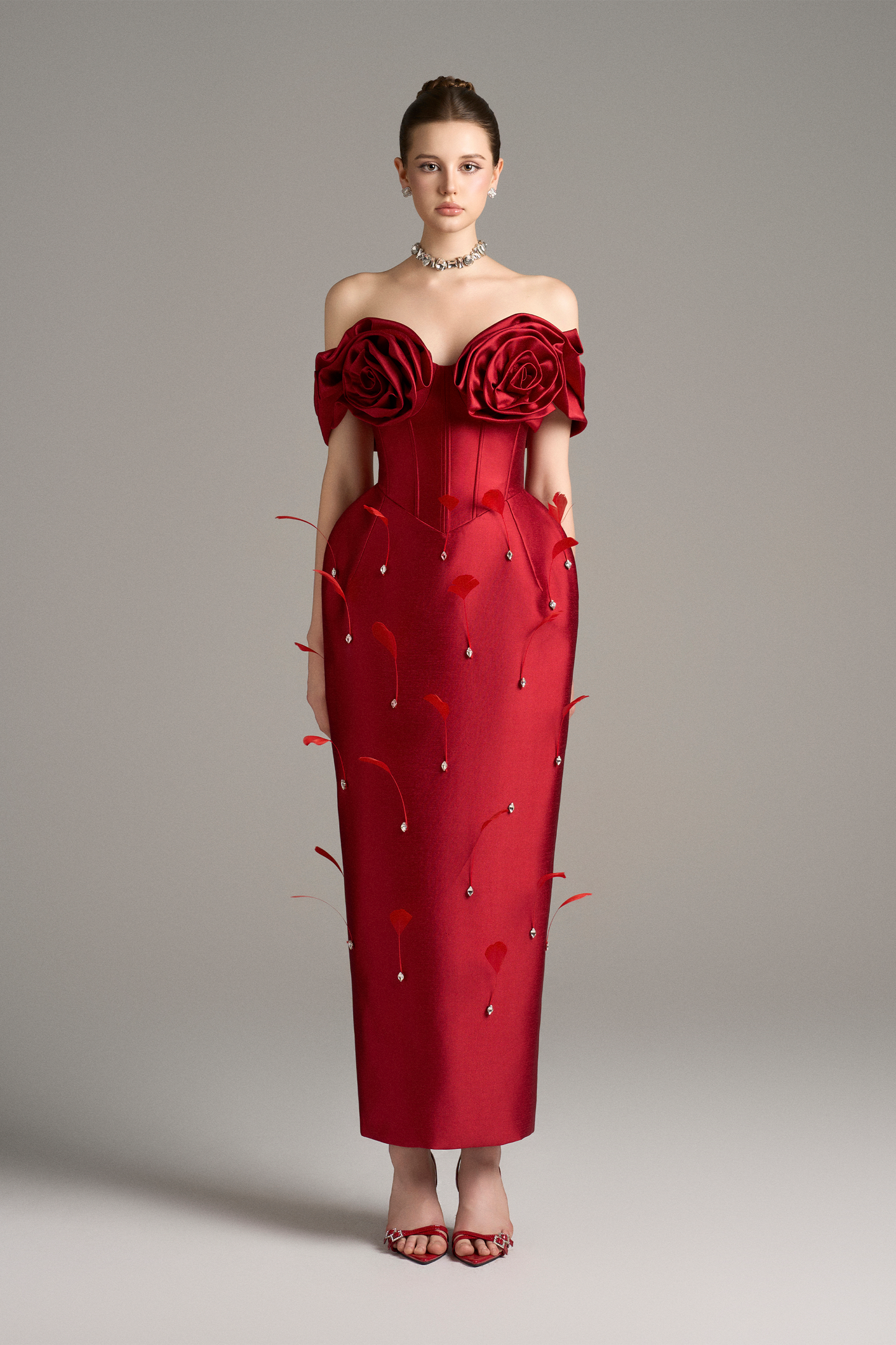 Full-body shot of a model wearing a red Peony off-shoulder maxi dress with 3D roses, a corset, crystal beads, and feathers, displaying its elegant silhouette and unique details.
