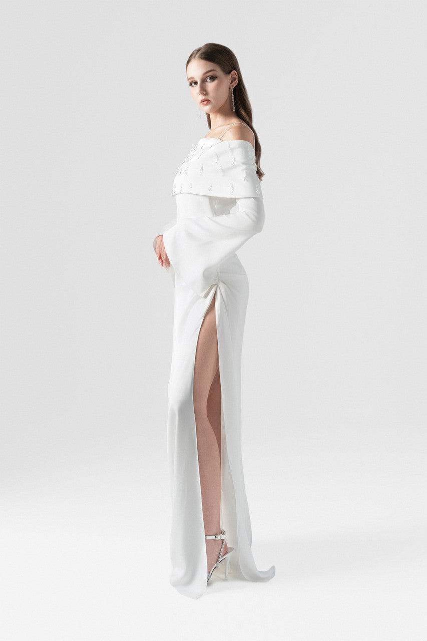 White maxi dress in silk with off-shoulder design, crystal embellishments, and a high side slit with pleating.