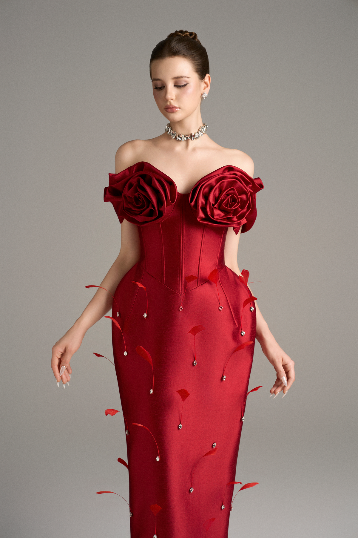 Detailed view of a red off-shoulder dress with oversized 3D petal roses, a fitted corset, and delicate crystal and feather embellishments, emphasizing the handcrafted elements.