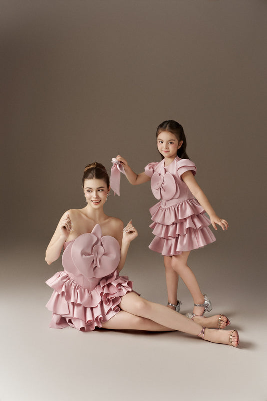 Two matching pink dresses, one adult and one child-sized. The adult dress is strapless with a 3D handmade flower bust detail and a ruffled skirt. The child's dress has layered puff sleeves, a round neckline, a 3D handmade flower bust detail, and a ruffled skirt.