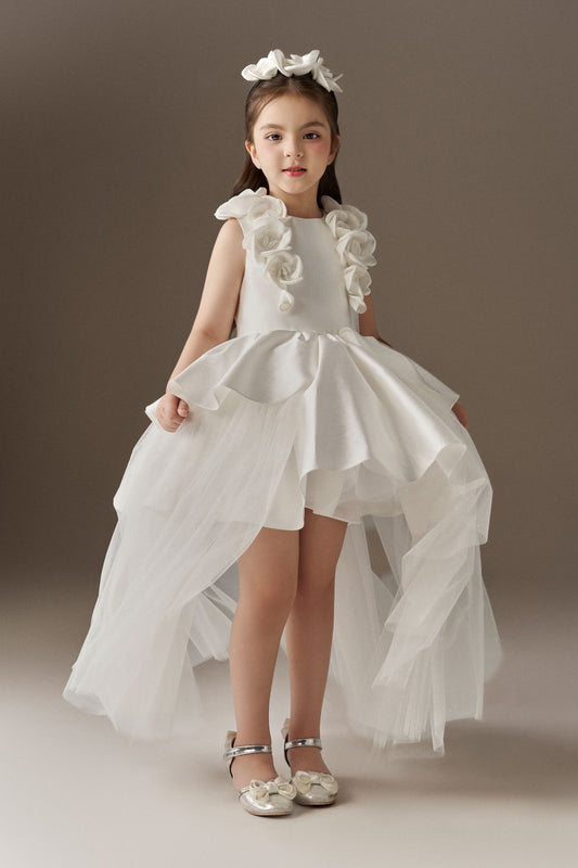 A white sleeveless princess dress with a fitted bodice and a round neckline, accented by handcrafted floral embellishments on the shoulders. The dress features a multi-layered skirt with an outer layer of glitter mesh tulle, designed with a high-low hem. A floral headband completes the look.