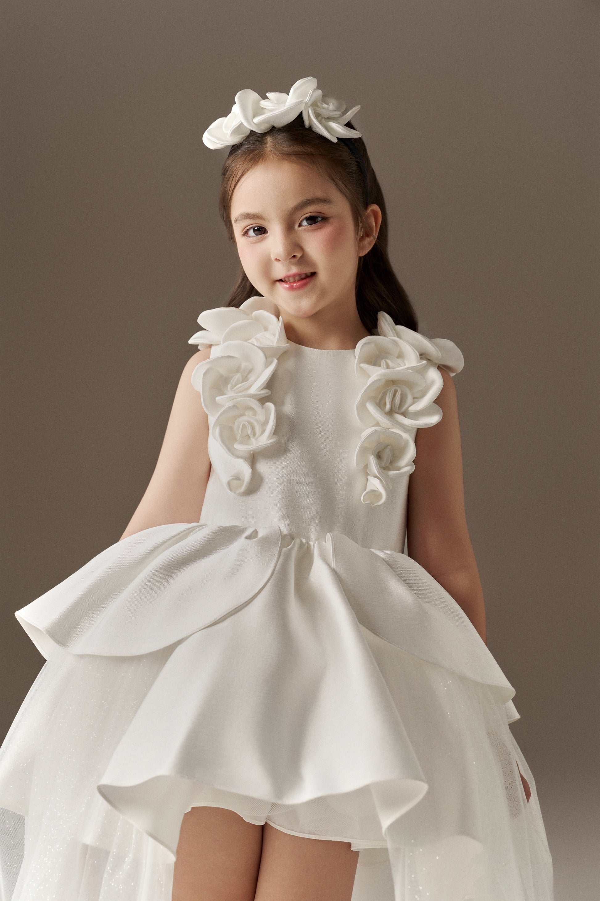A white sleeveless princess dress featuring a fitted bodice with a round neckline and handcrafted floral embellishments on the shoulders. The dress has a multi-layered skirt with an outer layer of glitter mesh tulle, creating a high-low effect. A floral headband is worn.