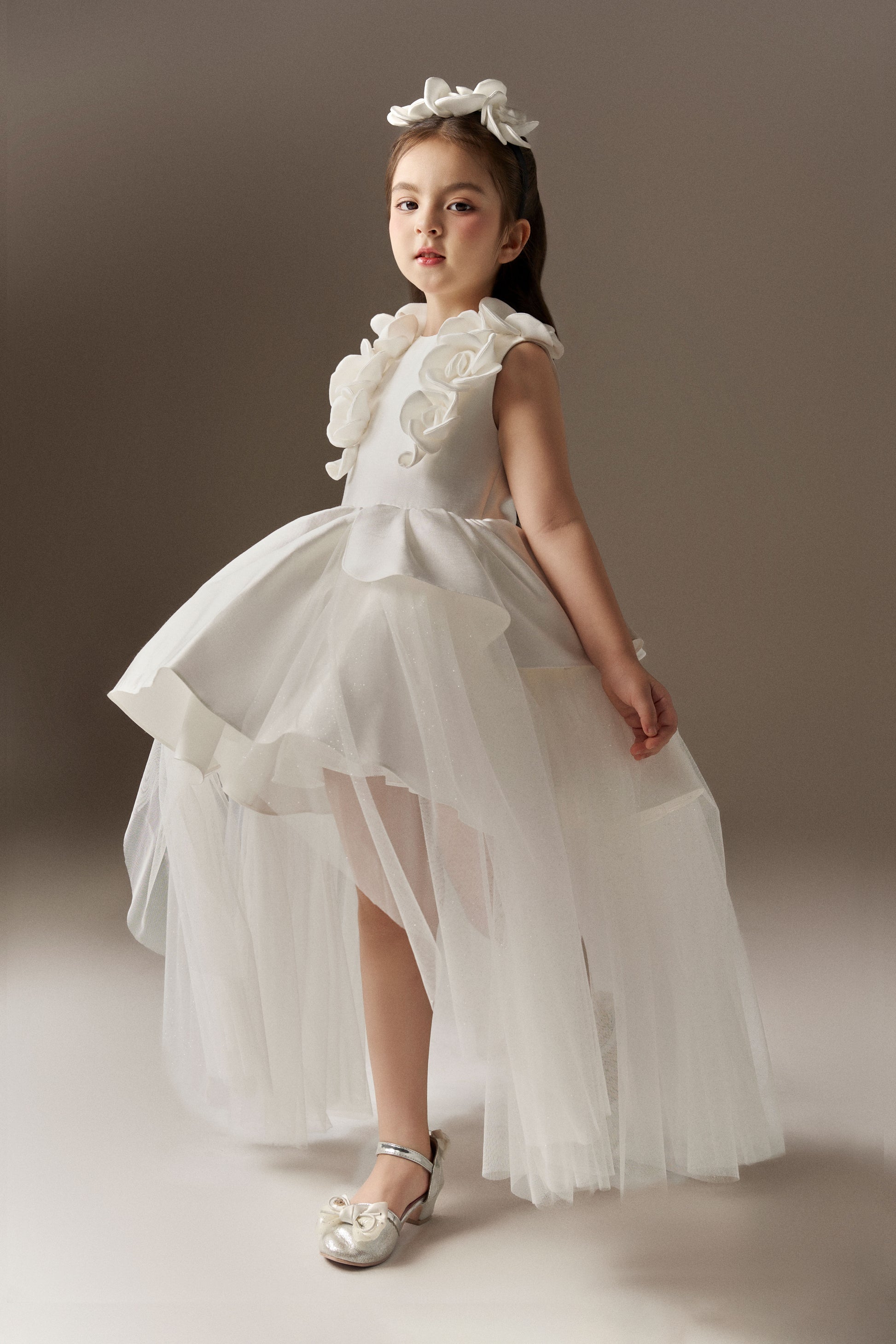 A white sleeveless princess dress with a fitted bodice and a round neckline, accented by handcrafted floral embellishments on the shoulders. The dress features a multi-layered skirt with an outer layer of glitter mesh tulle, designed with a high-low hem. A floral headband completes the look.