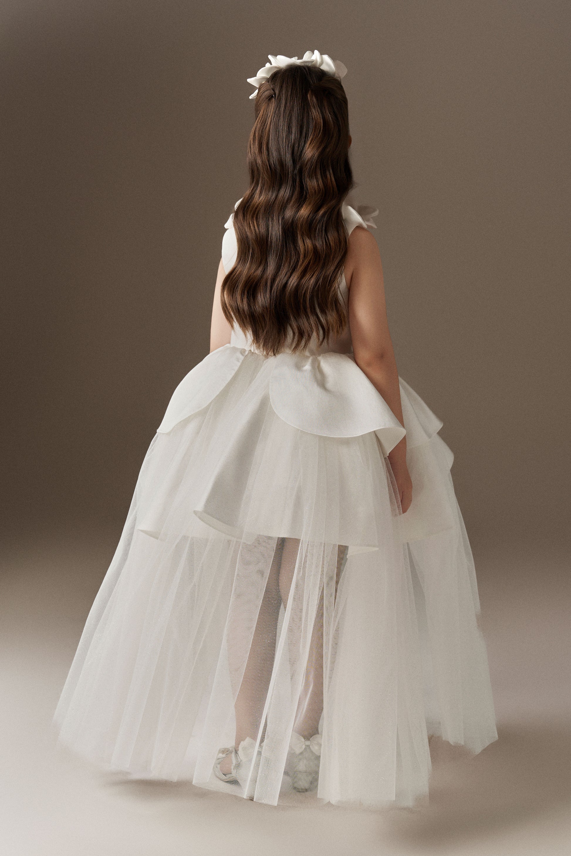 The back view of a white sleeveless princess dress, showcasing its voluminous multi-layered skirt with an outer layer of glitter mesh tulle and a high-low hem. Handcrafted floral embellishments are visible on the shoulders.