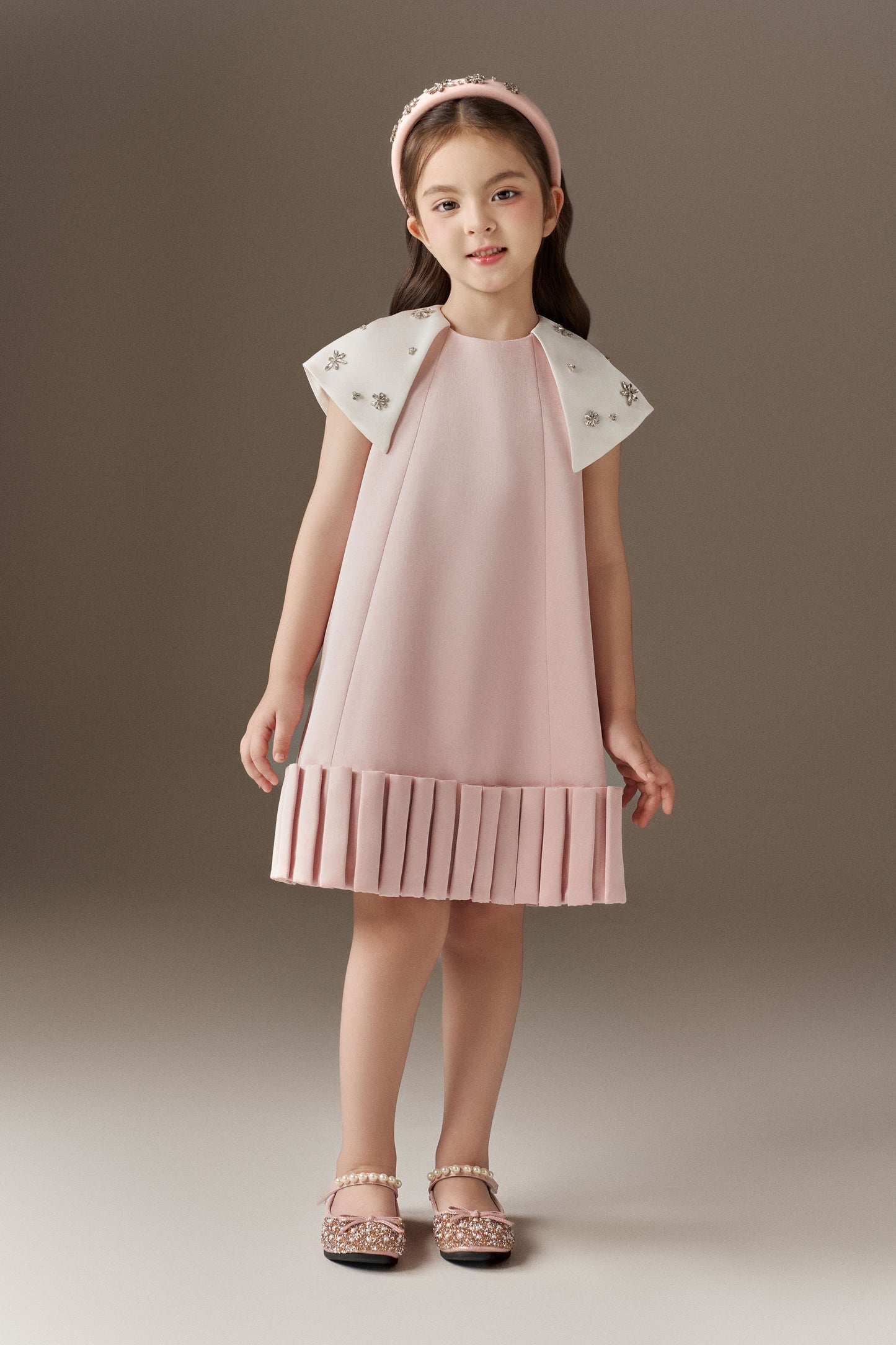 A pink A-line children's dress with a round neckline, featuring a distinctive 3D pleated hem and white detachable cape sleeves embellished with sparkling beads. The ensemble includes a pink rhinestone headband.