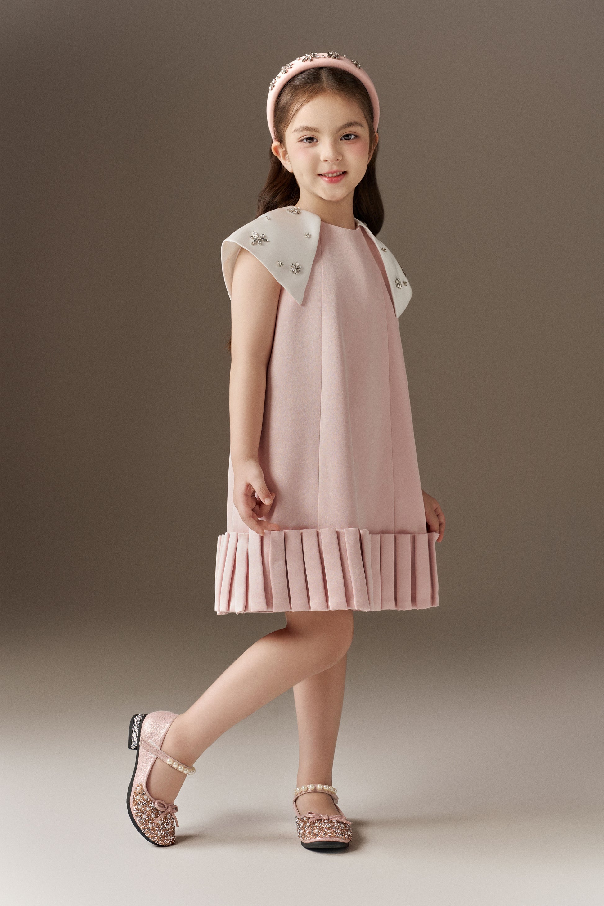 A pink A-line children's dress with a round neckline, a unique 3D pleated hem, and white detachable cape sleeves adorned with sparkling beads. A pink rhinestone headband is included.