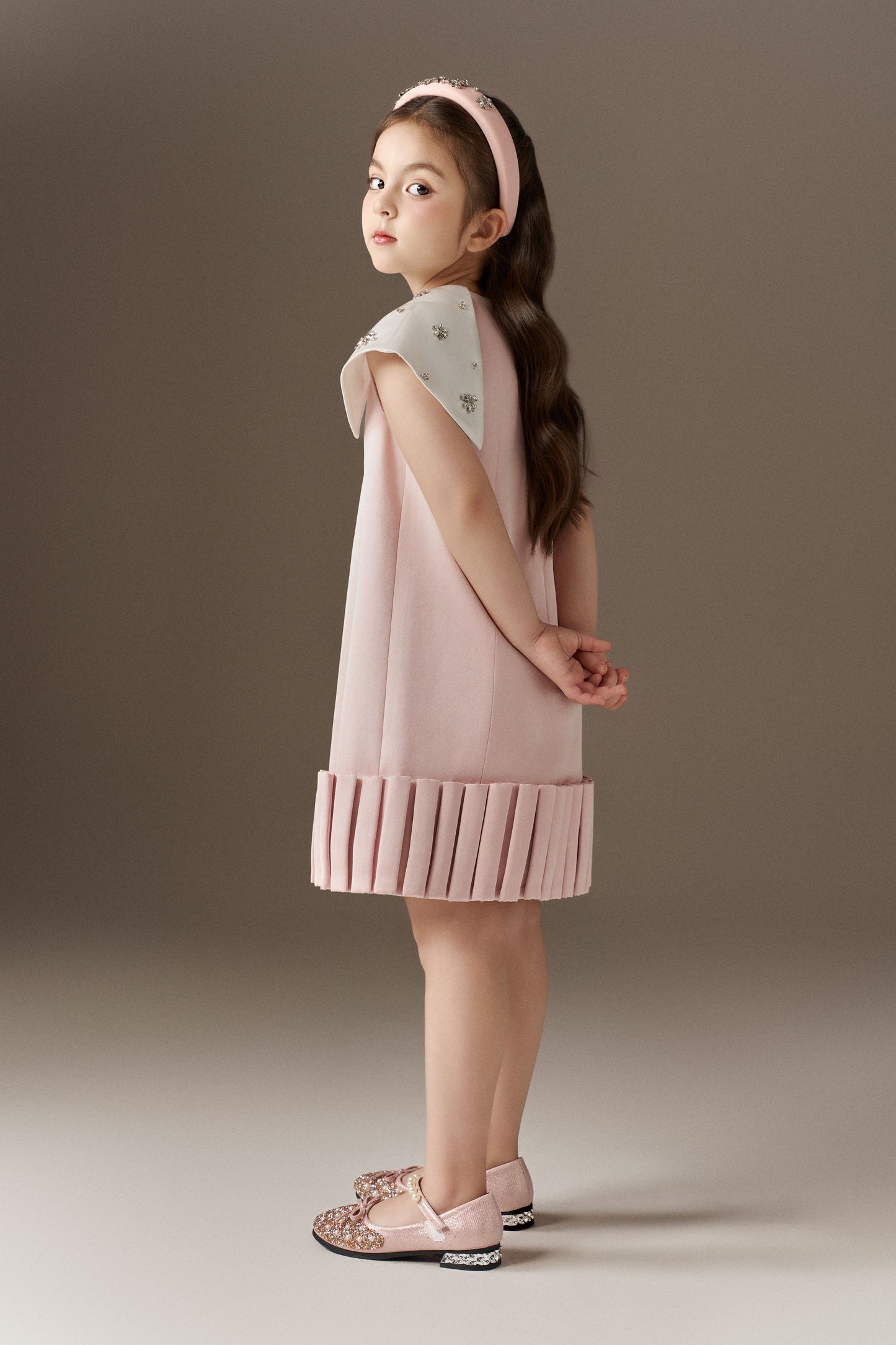 A pink A-line children's dress with a round neckline, featuring a 3D pleated hem effect and white detachable cape sleeves embellished with sparkling beads. A pink rhinestone headband is worn.