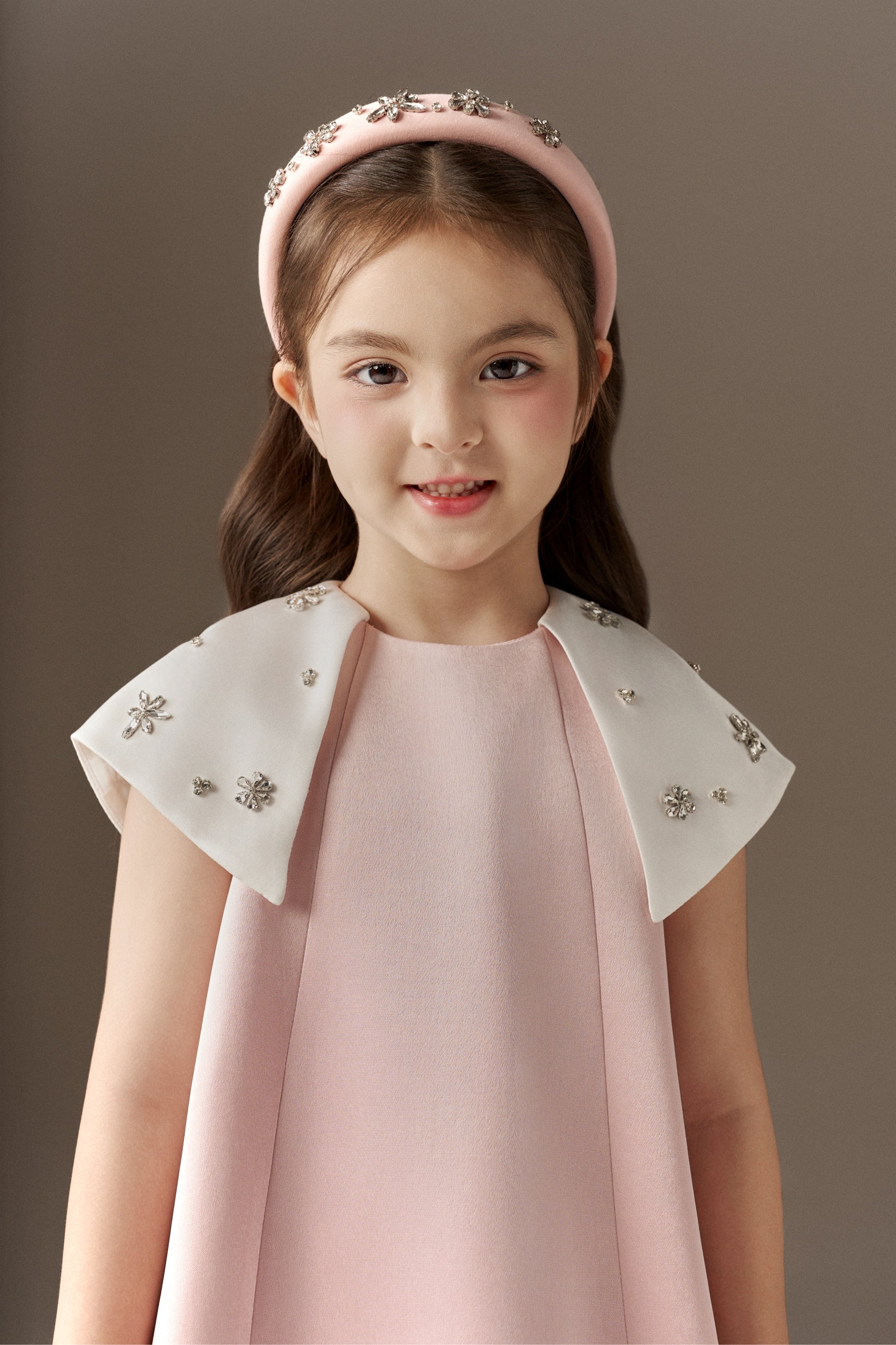 A close-up of a pink A-line children's dress, highlighting its round neckline and the white detachable cape sleeves embellished with sparkling beads. A pink rhinestone headband with intricate detailing is visible.
