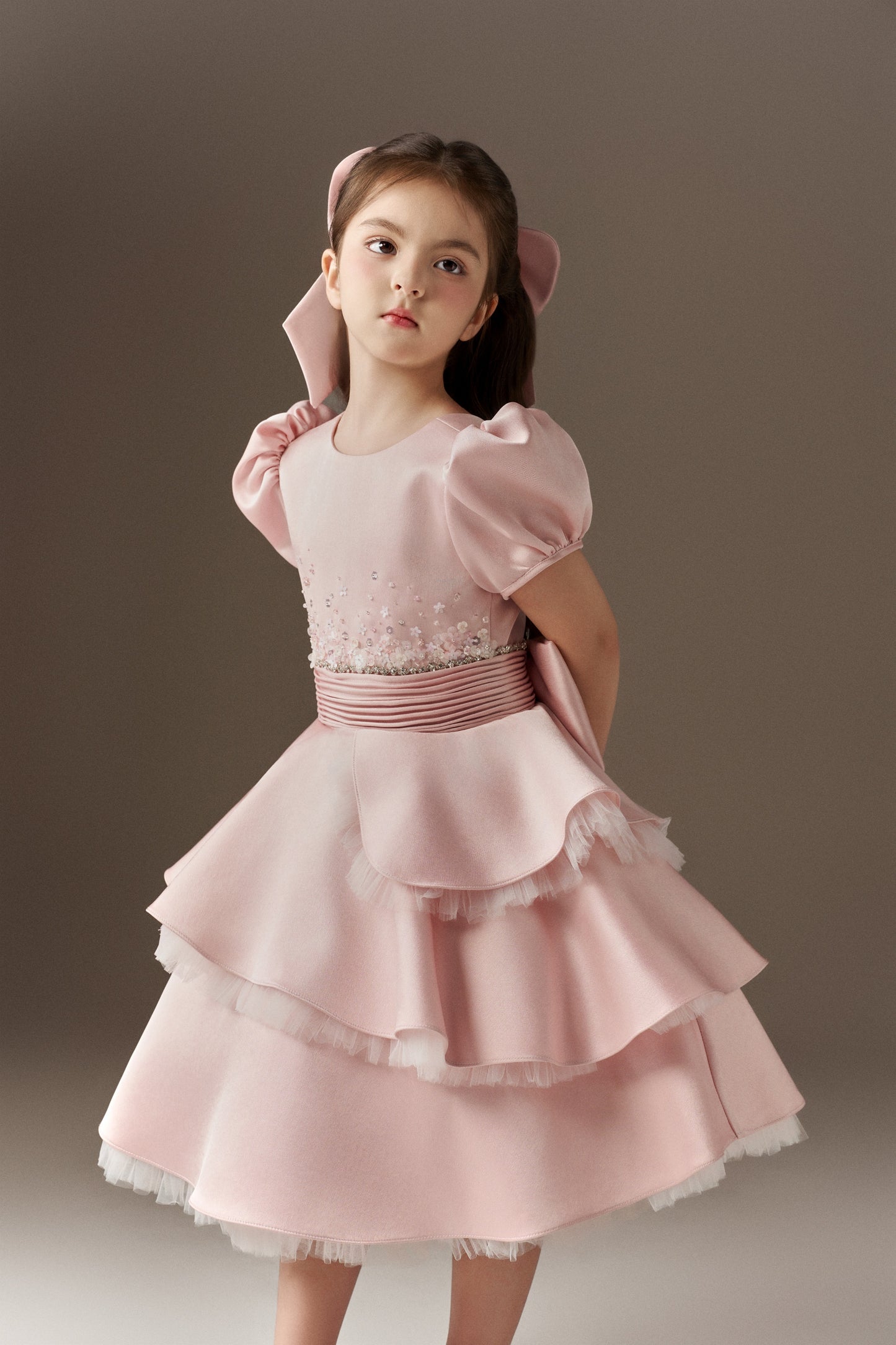 A pink children's dress showcasing a round neckline, puff short sleeves, and a fitted bodice decorated with hand-applied floral embellishments, sequins, crystals, and beads. The dress features a soft belted waist and a layered, ruffled skirt. An oversized pink hair bow is included.