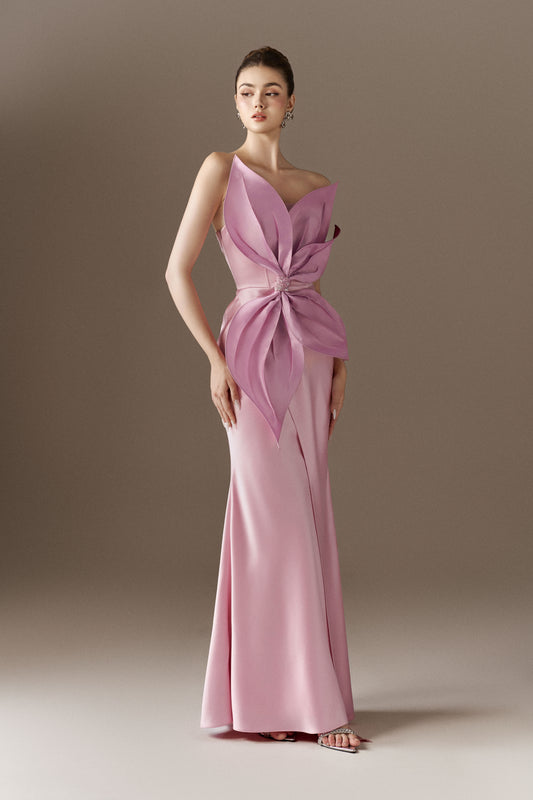 A pink, sleeveless, figure-hugging maxi dress, highlighting its bandeau neckline, off-shoulder open neckline, and the prominent 3D handcrafted petal detail at the waist. The dress also features a front slit.