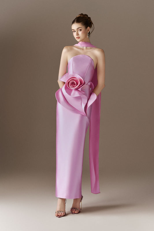 A close-up view of a pink strapless maxi dress, highlighting the bandeau neckline, the fitted bodice, and an elaborate, oversized 3D rose embellishment on the left hip, with a long, narrow scarf draped over the right shoulder.