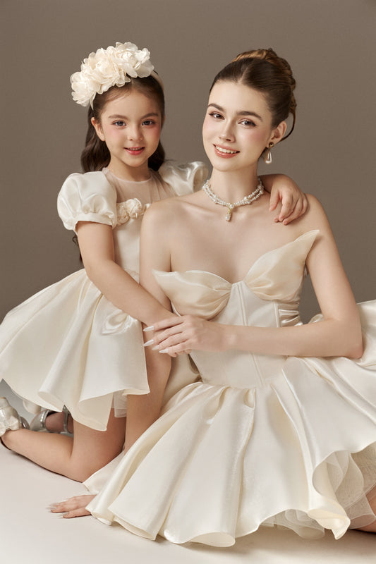 Two matching ivory dresses, an adult and a child's version. The adult dress features a strapless sweetheart neckline and a voluminous puffed skirt. The child's dress has short puff sleeves, a sheer mesh neckline, a fitted bodice, and a voluminous puffed skirt. A floral headband is worn by the child.