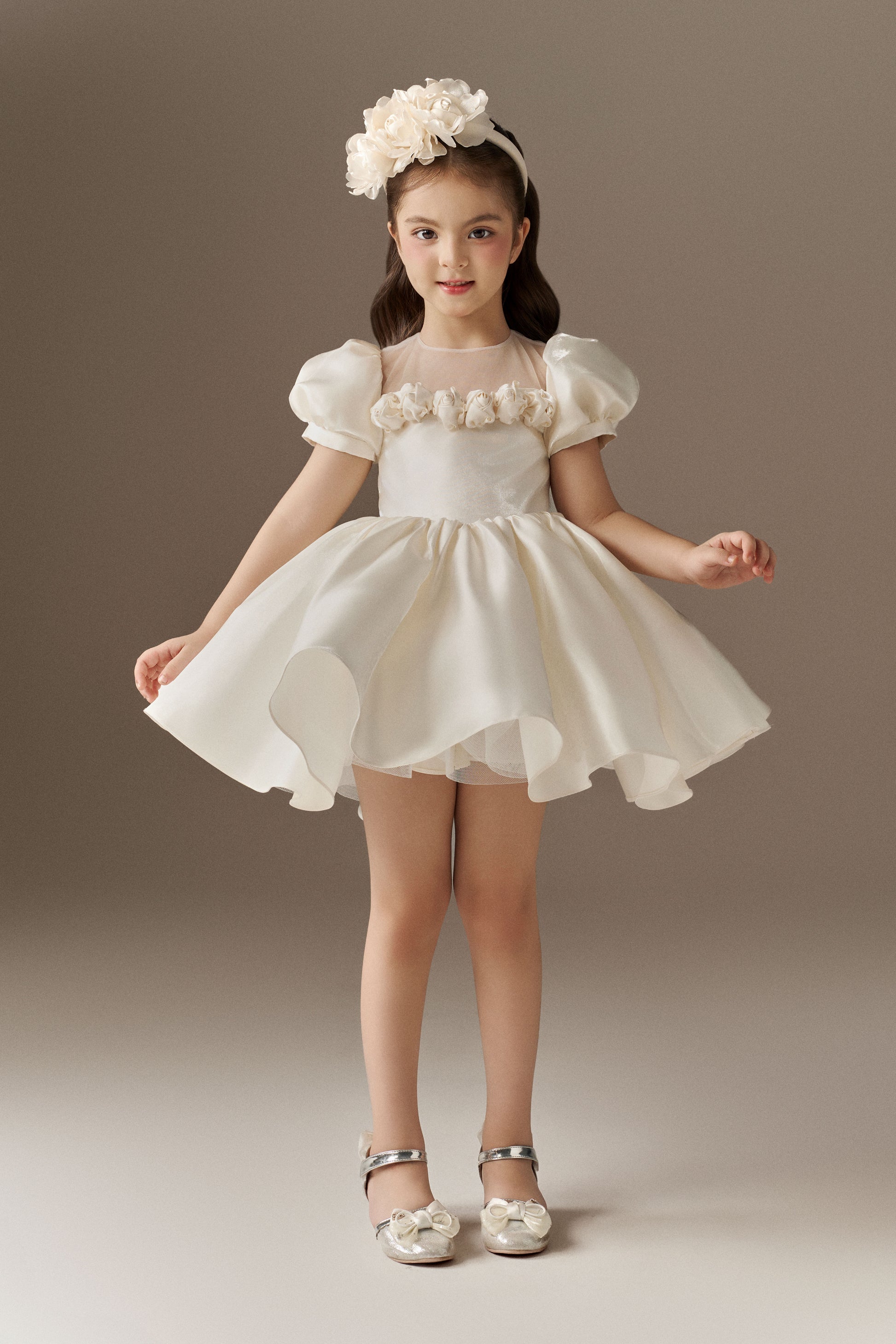 An ivory child's dress with short puff sleeves and a sheer mesh neckline. The bodice is fitted, and the voluminous puffed skirt is adorned with hand-sewn floral embellishments. A floral headband is included.