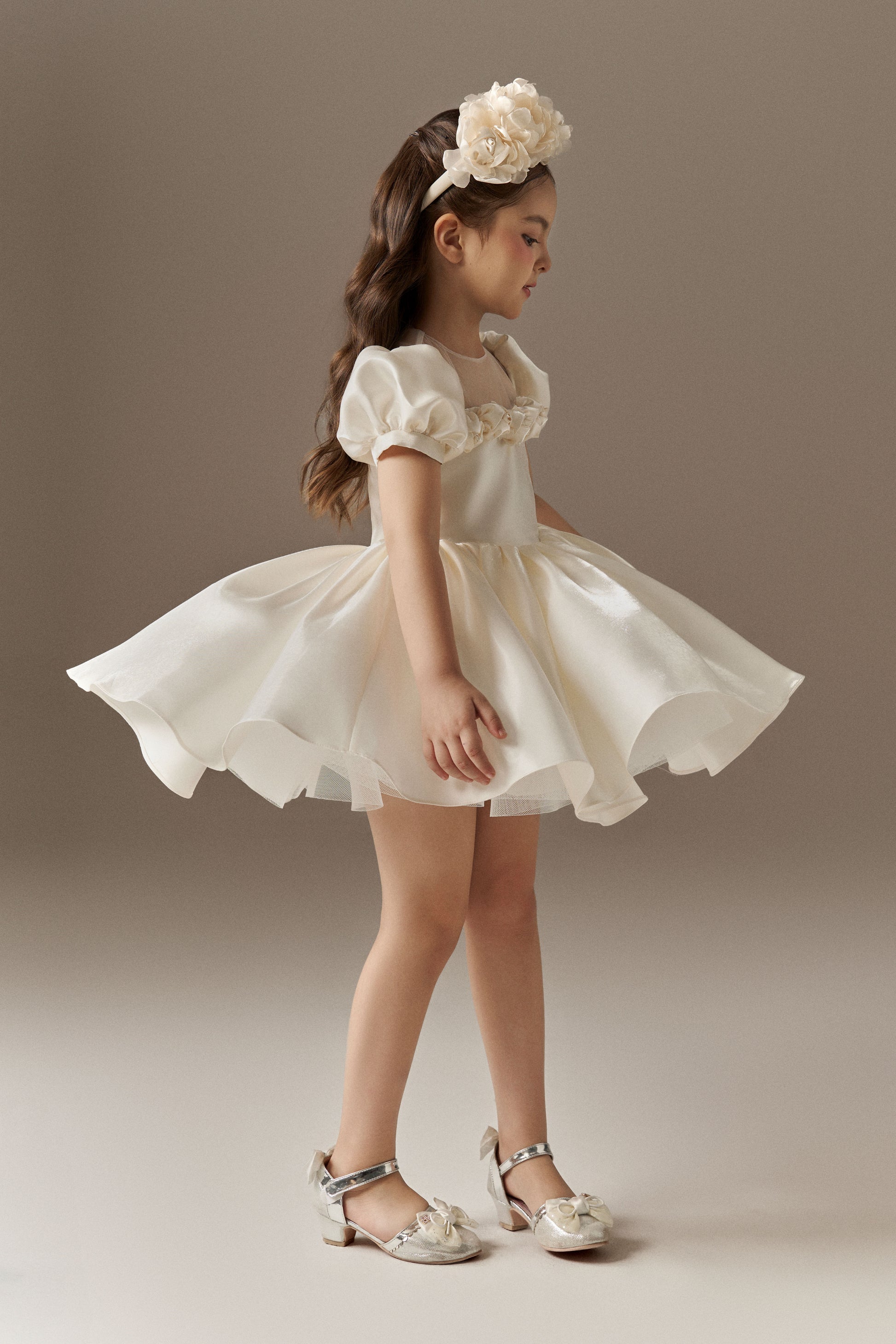 An ivory child's dress with short puff sleeves and a sheer mesh neckline. The bodice is fitted, and the dress features a voluminous puffed skirt with hand-sewn floral embellishments. A floral headband is worn.