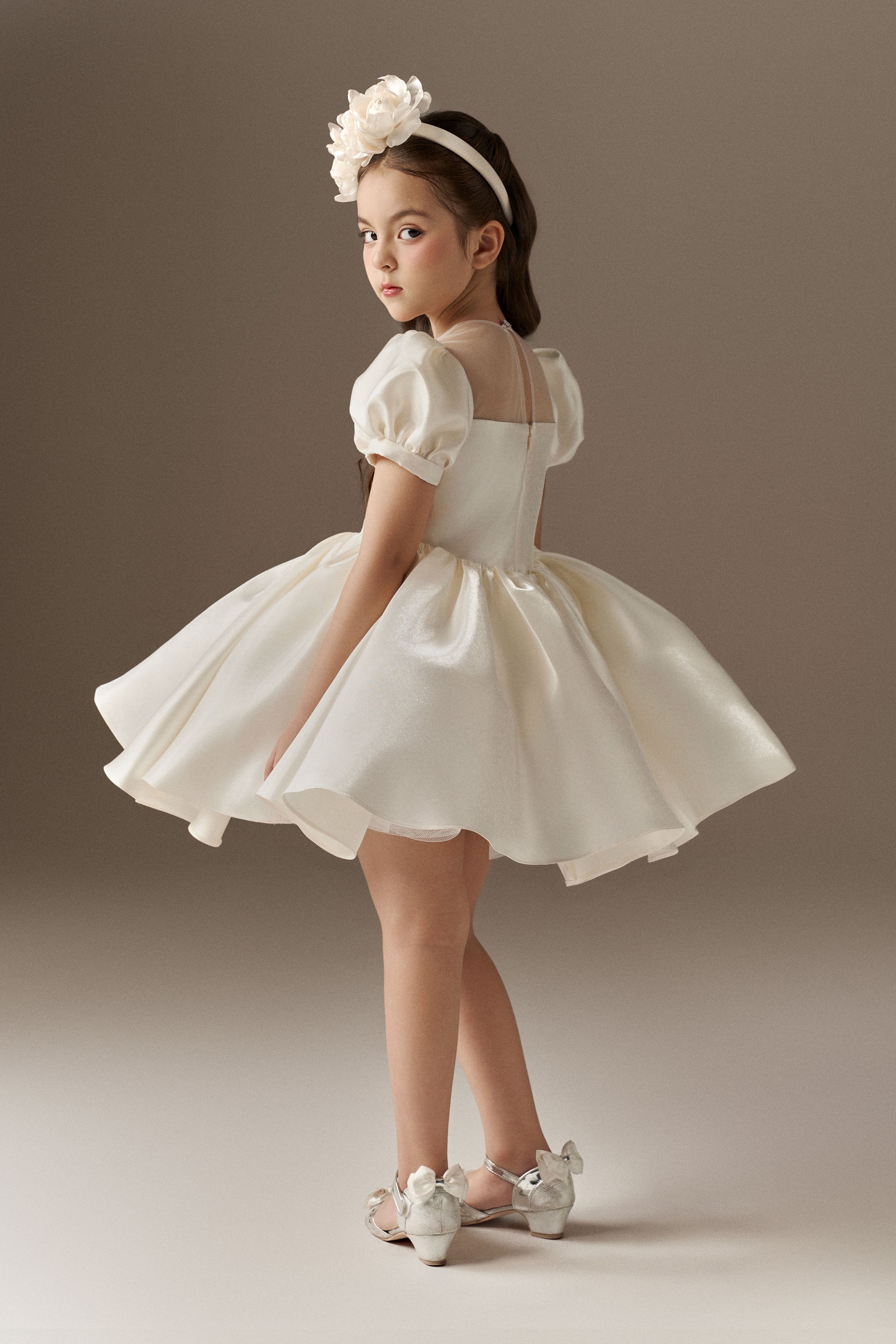 A side view of an ivory child's dress with short puff sleeves and a sheer mesh neckline. The dress features a voluminous puffed skirt with hand-sewn floral embellishments. A floral headband is worn.