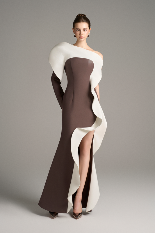 Model wearing a brown floor-length gown with a dramatic white sculptural ruffle cascading from the shoulder down the front, featuring an asymmetrical neckline and thigh-high slit.