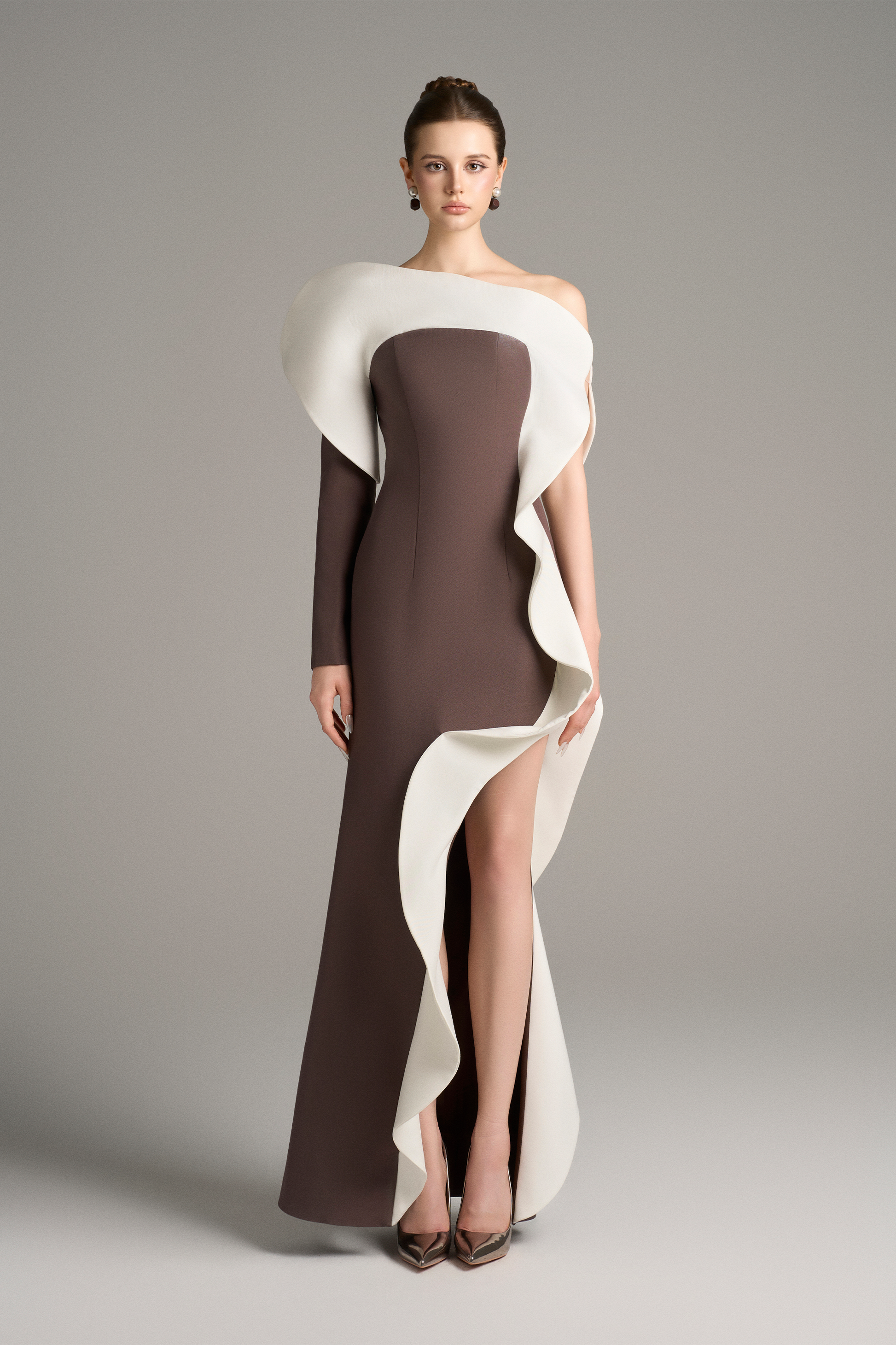 Model wearing a brown floor-length gown with a dramatic white sculptural ruffle cascading from the shoulder down the front, featuring an asymmetrical neckline and thigh-high slit.
