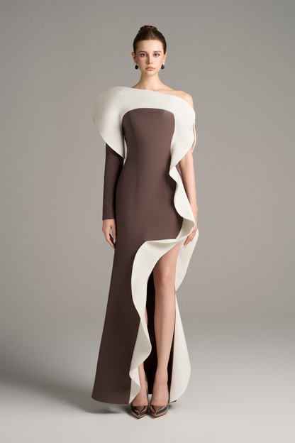 Model wearing a brown floor-length gown with a dramatic white sculptural ruffle cascading from the shoulder down the front, featuring an asymmetrical neckline and thigh-high slit.