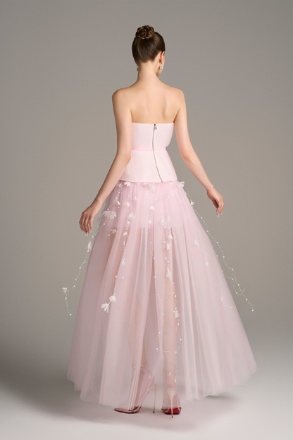 Back view of Aveta bandeau top maxi dress in pastel pink featuring corset bodice, copper zipper, and flowing sheer layered skirt with floral appliqué details