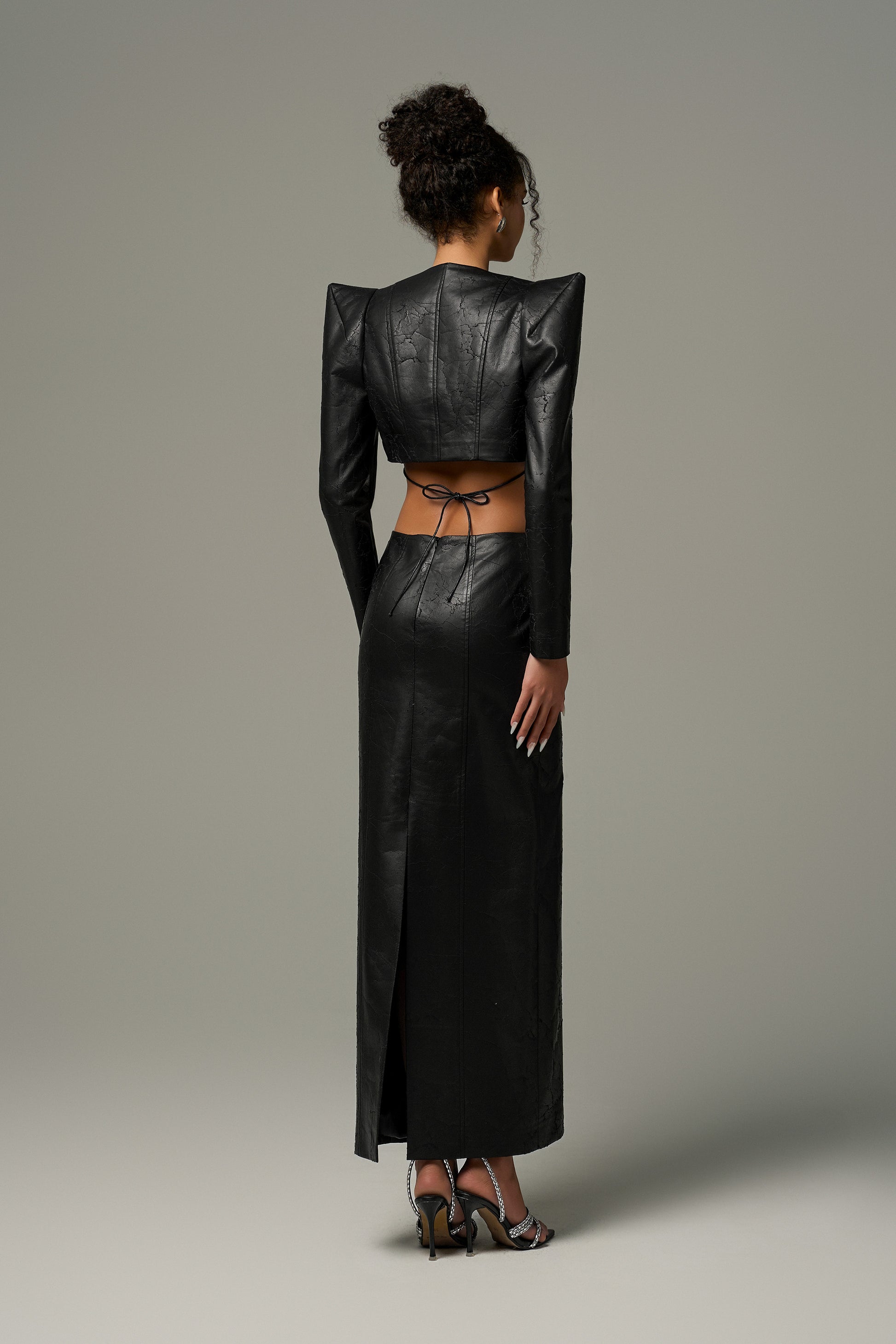 Black leather ensemble features a cropped jacket with sharp shoulders and a form-fitting skirt with a daring back slit and lace-up details.