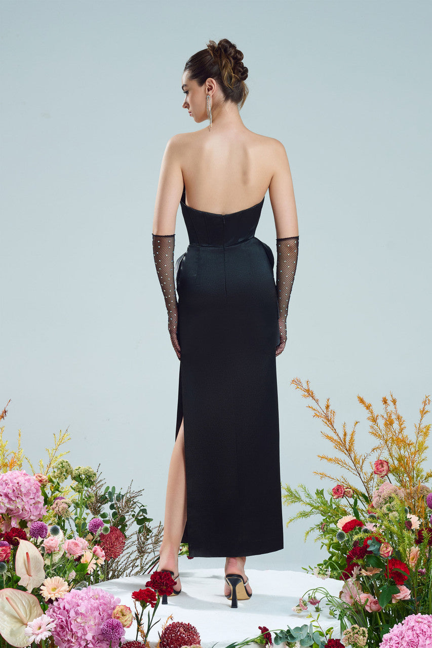 A black dress, featuring a bold side slit, a crystal-embellished neckline, and asymmetrical draping at the waist.