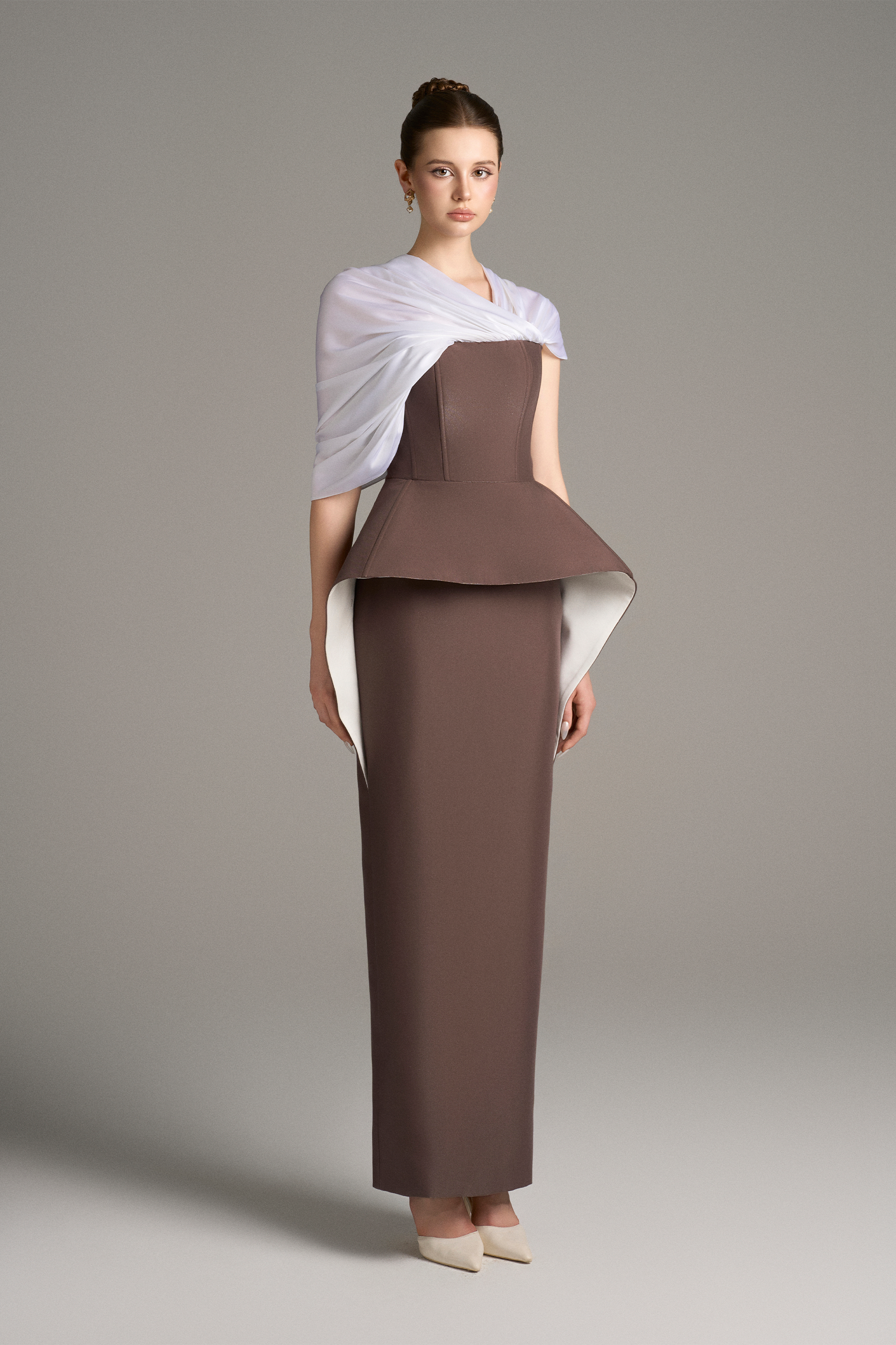 Three-quarter view of strapless column gown with structured peplum detail and draped white shoulder wrap