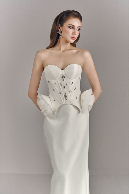 Woman wearing a strapless white gown with intricate detailing and white gloves against a plain background