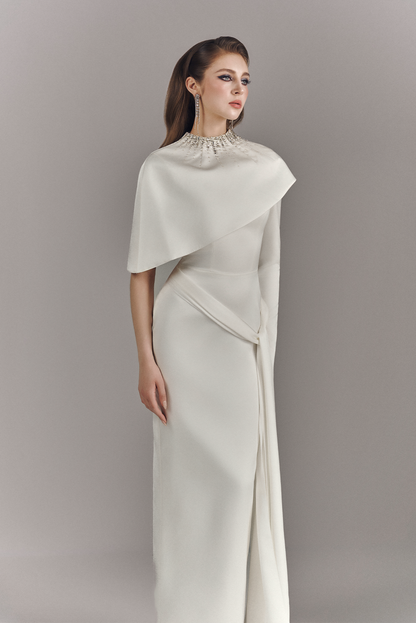 Woman wearing a long, elegant white dress with a cape against a plain background
