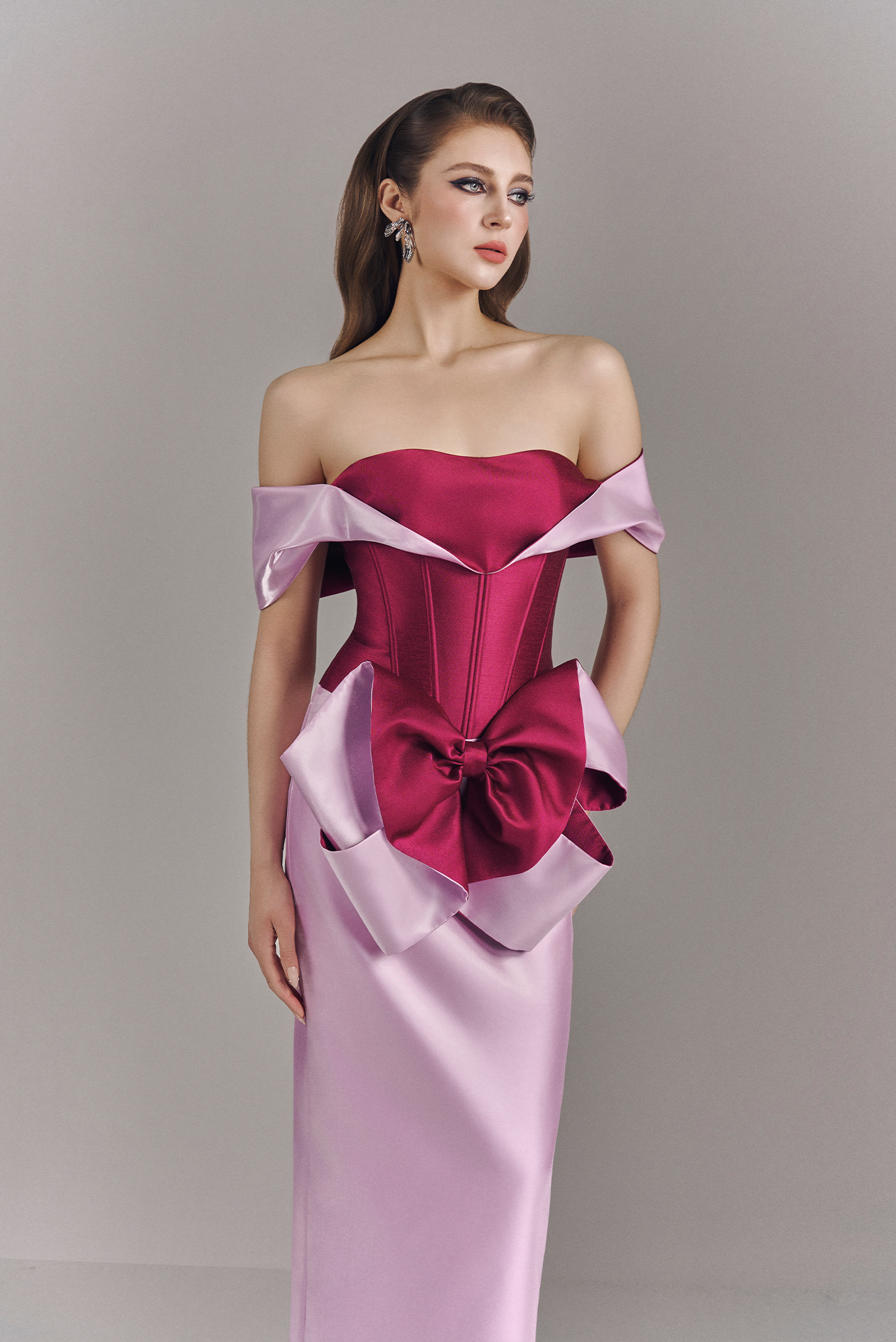 Woman wearing a pink off-shoulder gown with a large bow detail on a gray background