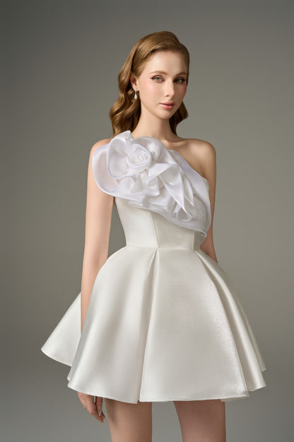 A close-up front view of a graceful white flared micro-mini dress, highlighting the bandeau neckline, the prominent one-shoulder strap with handcrafted floral embellishments, and the corset bodice.