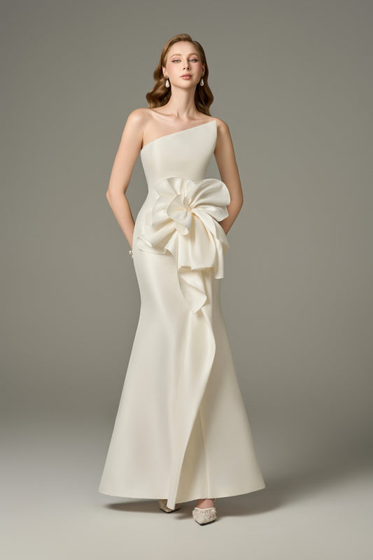 A full-length front view of a Devotion ivory acetate maxi dress with a fit-and-flare, figure-hugging silhouette, featuring an asymmetric bandeau neckline with a dramatic 3D oversized handcrafted floral embellishment on one shoulder.