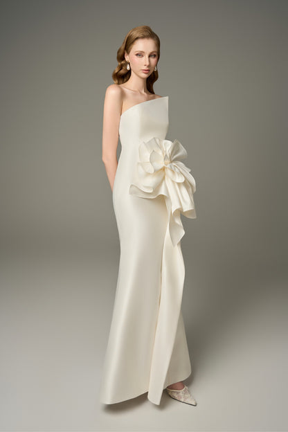  A side-angle view of a Devotion ivory acetate maxi dress, emphasizing its fit-and-flare silhouette and the striking 3D oversized handcrafted floral embellishment on the asymmetric neckline.