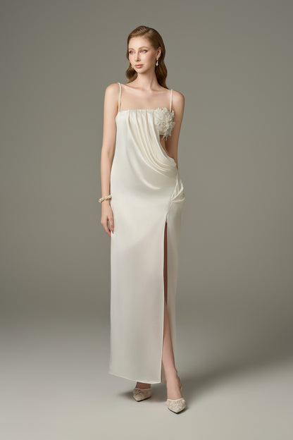  A white, figure-hhugging maxi dress featuring a bandeau neckline with spaghetti straps, a thigh-high slit, draped details on the side, and handcrafted floral embellishments on the bodice.