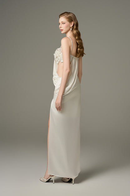 A back and side view of a white, sleeveless maxi dress with a thigh-high slit, showcasing its figure-hugging fit, draped details, and floral embellishments on the side of the bodice.