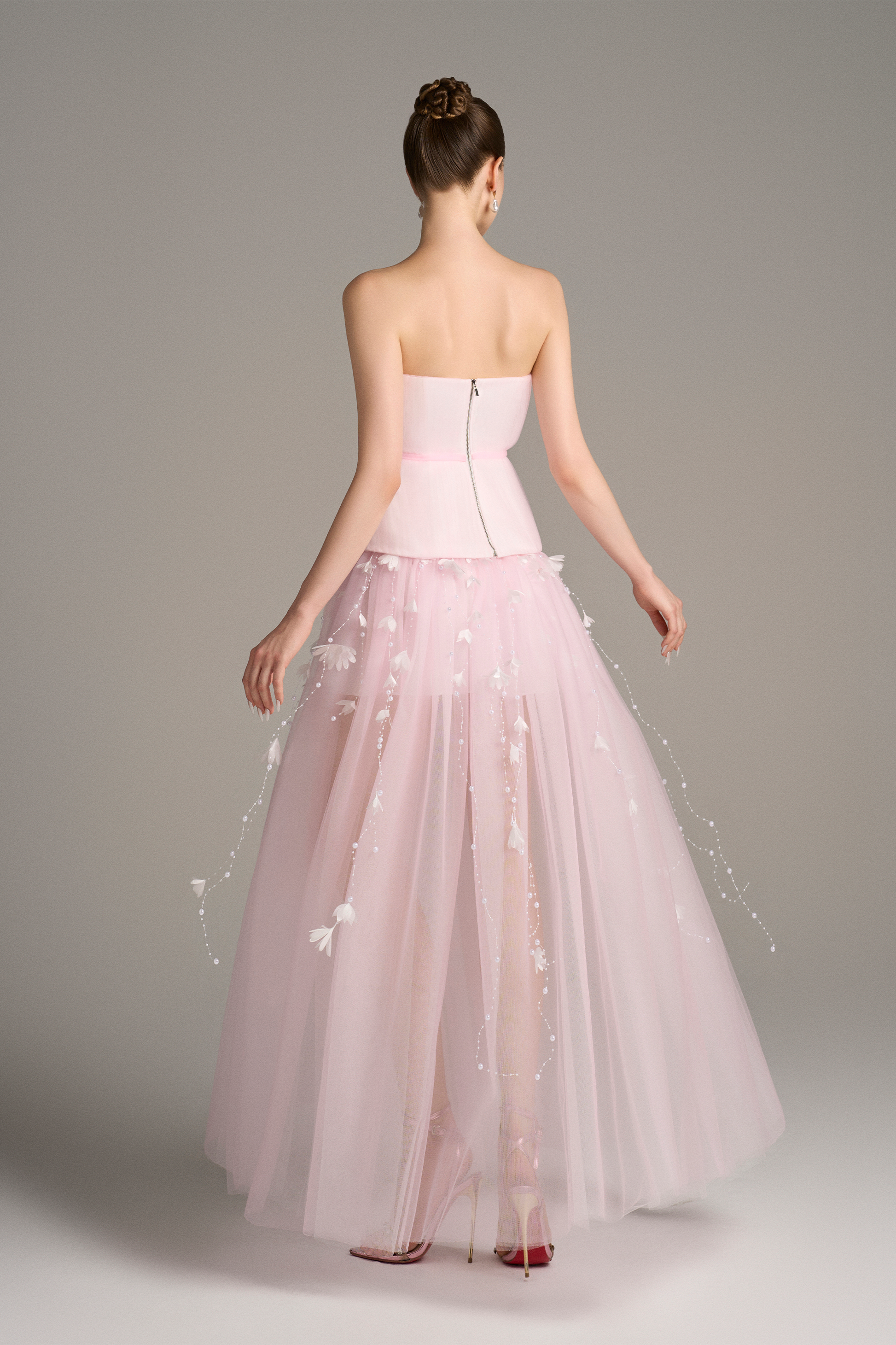 Back view of Aveta bandeau top maxi dress in pastel pink featuring corset bodice, copper zipper, and flowing sheer layered skirt with floral appliqué details