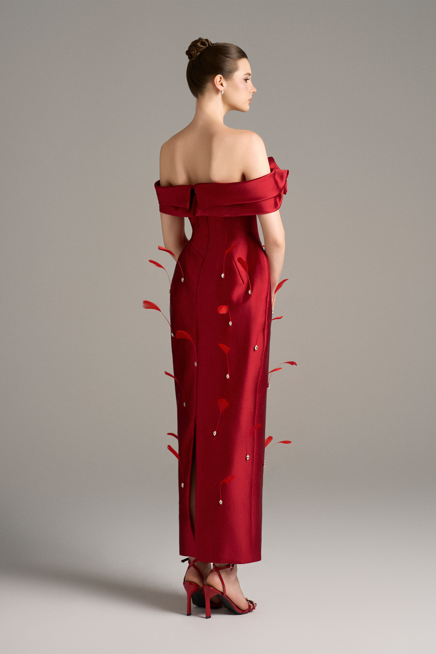 Back view of a red off-shoulder maxi dress featuring a corset, 3D rose details, feather accents, and a back slit, highlighting the dress's structure and embellishments.