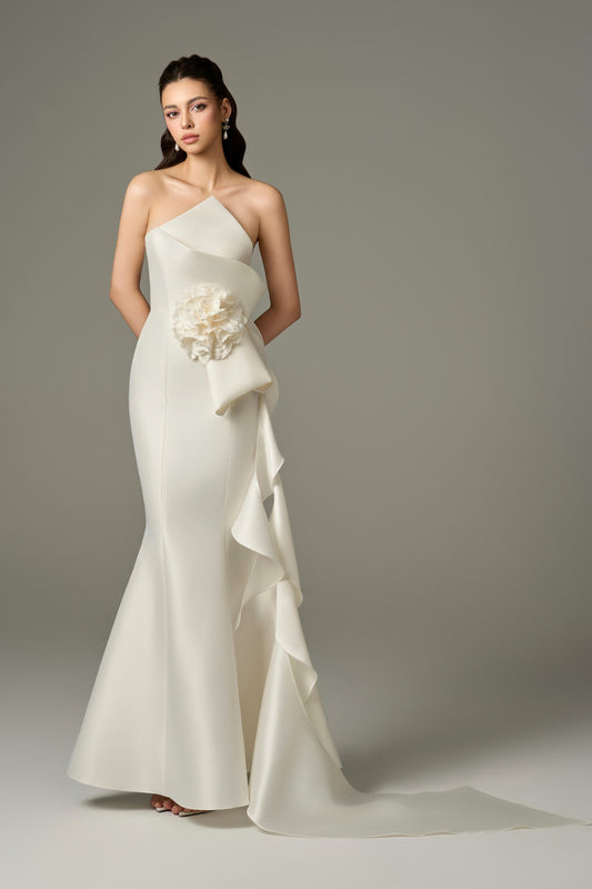 A full-length front view of an Isabella ivory acetate fishtail gown with a figure-hugging fit, featuring a bandeau neckline, sleeveless design, draped details on the side, and a large, handcrafted 3D floral embellishment on the hip.
