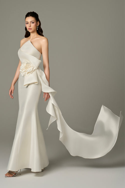 A side view of an Isabella ivory acetate fishtail gown, emphasizing the draped details, the figure-hugging fit of the bodice, and the dramatic flair of the fishtail skirt with the large 3D flower.