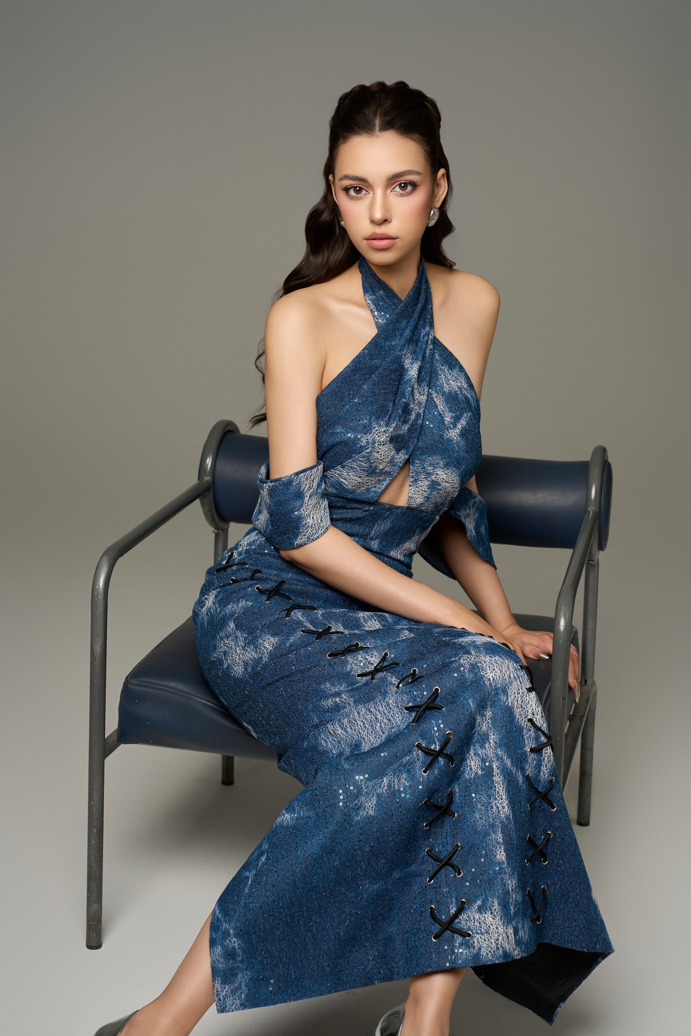A seated view of a chic modest blue cotton denim halter tie-dye maxi dress, emphasizing the halter neckline, off-shoulder design, and the front criss-cross black laces on the dress.
