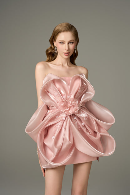 A close-up front view of a pink, sleeveless mini dress with a bandeau neckline, emphasizing the large, handcrafted "petal" flower detail at the front of the bodycon dress.