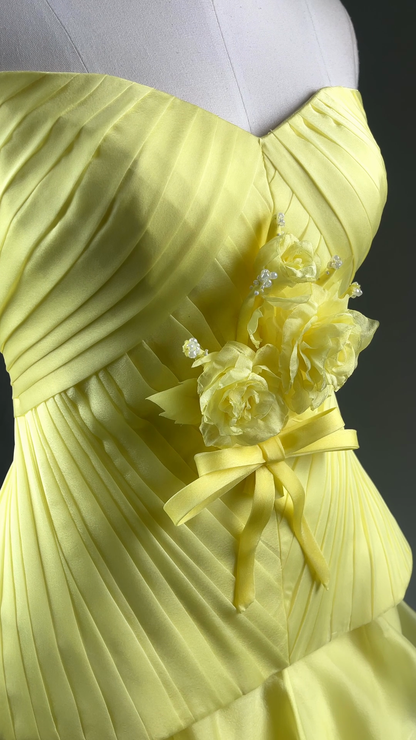 A close-up of a pale yellow strapless gown on a white mannequin torso, featuring pleated fabric, a sweetheart neckline, and a floral embellishment with a yellow ribbon bow at the waist.