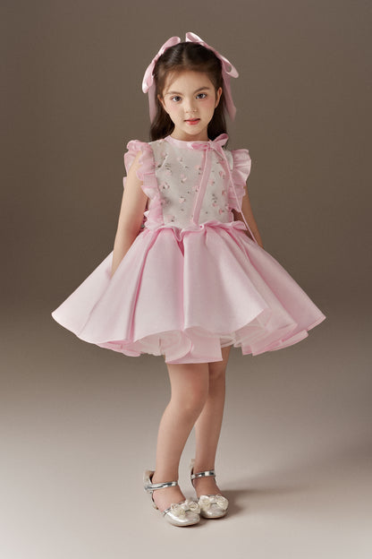 A pink sleeveless children's dress with a round neckline and prominent ruffle details on the front. The bodice is adorned with hand-applied beads and sequins, and the dress features a voluminous skirt. Two oversized pink hair bows are included.