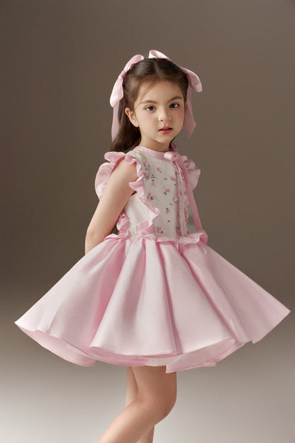A pink sleeveless children's dress with a round neckline, featuring large ruffle details on the front and a voluminous skirt. The bodice is embellished with hand-applied beads and sequins. Two oversized pink hair bows complete the look.