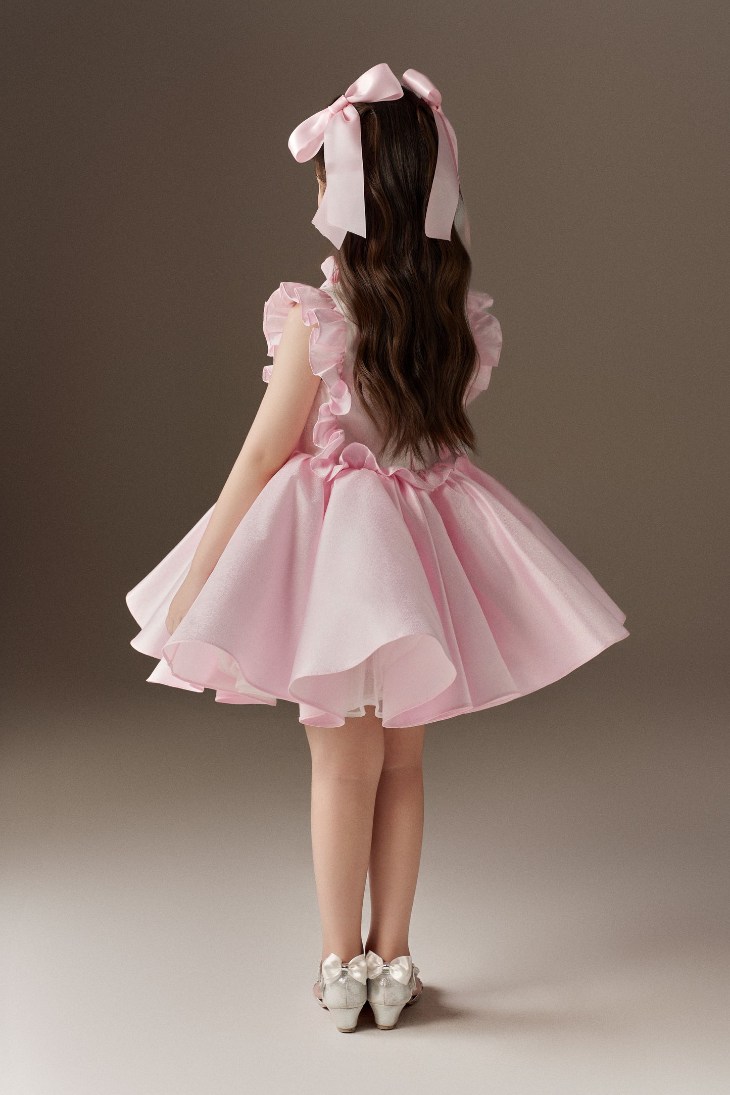 The back view of a pink sleeveless children's dress, highlighting its large ruffle details on the back and a voluminous skirt. Two oversized pink hair bows are visible in the hair.