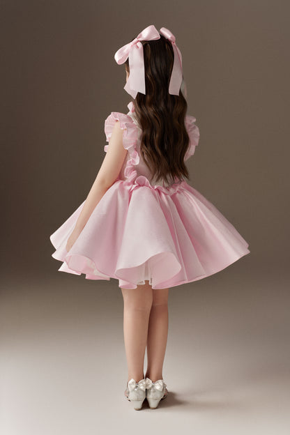 The back view of a pink sleeveless children's dress, highlighting its large ruffle details on the back and a voluminous skirt. Two oversized pink hair bows are visible in the hair.